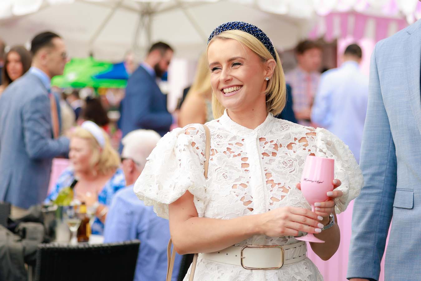 A blonde woman in a white lace dress holds a wine glass amid a crowd of people at a racecourse
