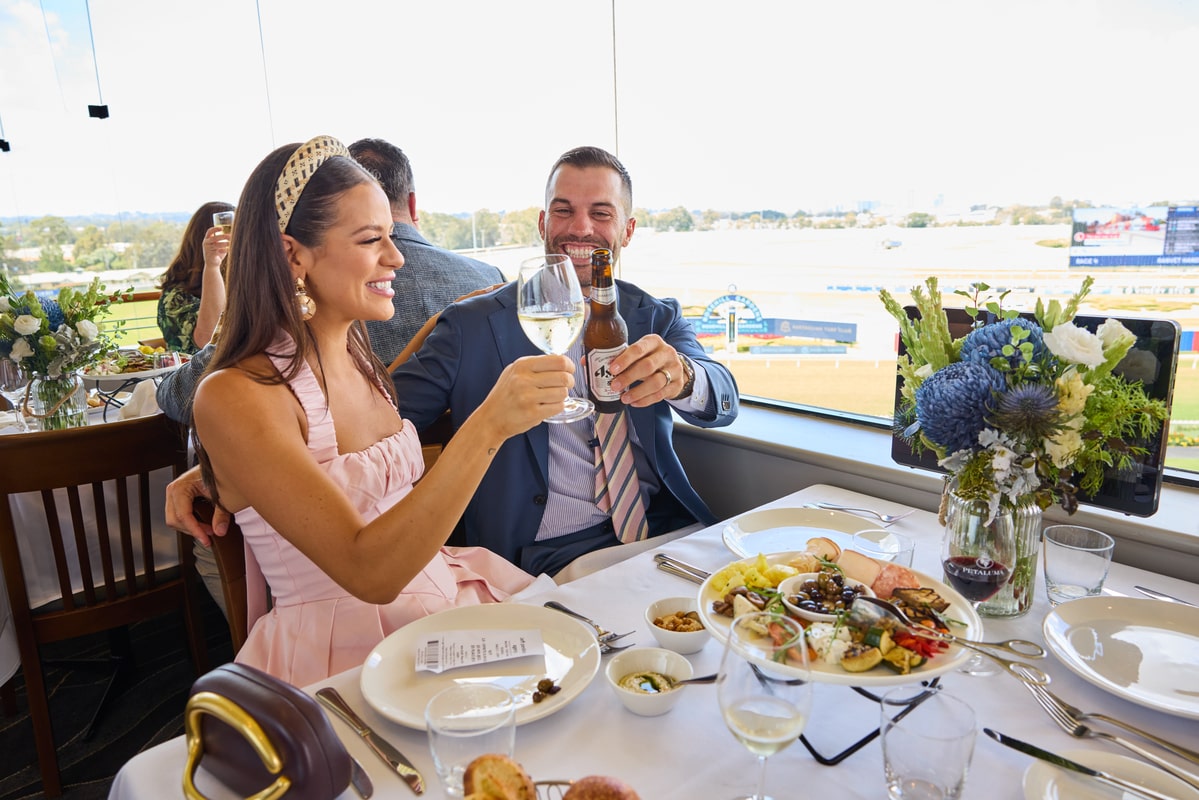 A man and woman cheers each other as they sit in a restaurant overlooking a racecourse